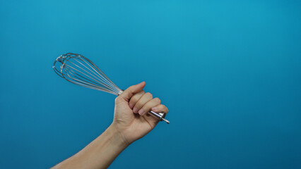 Caucasian man holding metal whisk isolated against blue wall background showcasing kitchen tool and cooking concept with simplicity and focus.