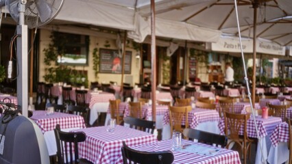 Outdoor cafe terrace with red gingham tablecloths, wooden chairs, umbrellas and sunlit patio, softly blurred bokeh cafe; background backplate copyspace calm.