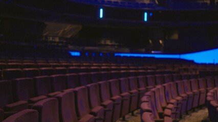 Obraz premium Blurred theater seating captured in shallow bokeh interior with curved rows, dim stage glow and soft purple seats suggesting an empty auditorium; backdrop copyspace overlay template.