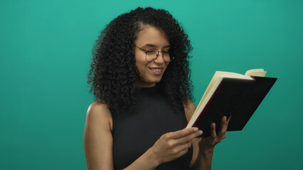 Young hispanic woman reading book over isolated green background wall looking amused and thoughtful.