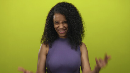 Young woman with curly hair and raised arms shows rock horns gesture in a yellow studio setting; fun vibe.