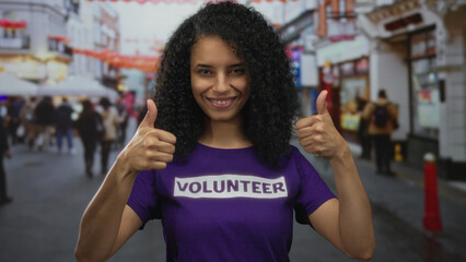 Latin woman points at purple volunteer shirt with thumbs up on busy city street during charity drive; pride duty.