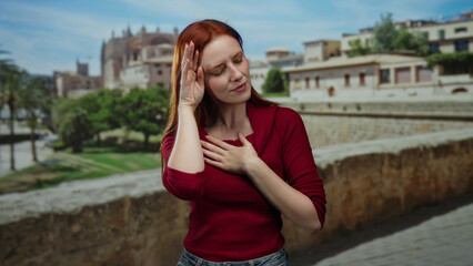 Fototapeta premium Woman standing on city street touches her head while wearing red sweater, surrounded by ancient buildings under clear sky, showcasing serene emotion and vibrant hair.