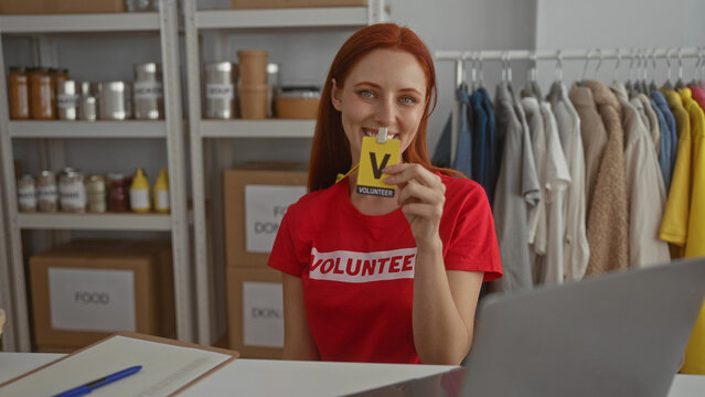 Woman volunteering indoors holding a volunteer badge in a donation room surrounded by food donation boxes and clothing in a charity center emphasizing redhead woman participation.