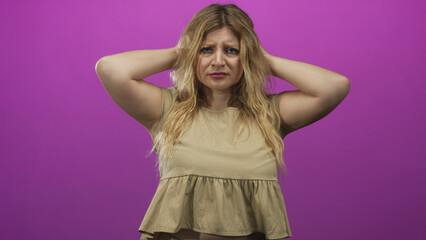 Young blonde woman holding head with hands, bare arms visible, tense worried facial expression in purple studio set; anxiety concern unease.