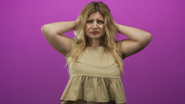 Young blonde woman holding head with hands, bare arms visible, tense worried facial expression in purple studio set; anxiety concern unease.