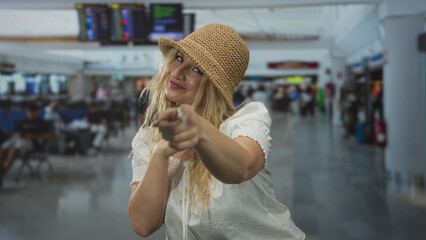Woman points finger at camera in airport terminal wearing straw hat and white blouse, forming a heart with hands in a playful pose; affection greeting travel.