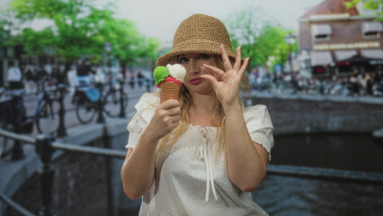 Woman holding ice cream cone by an amsterdam canal on a busy street making ok sign with hand and...