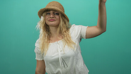 Woman with straw hat and blonde hair raises right arm in studio, wearing white peasant blouse and visible hand gesture against teal backdrop; playful confidence.