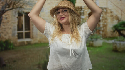 Woman blonde in straw hat raises both arms above her head while smiling in front of a sunlit stone building entrance wearing a white blouse; carefree joy.