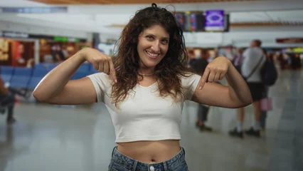 Fotobehang Beren Young woman points both fingers to bare abdomen in busy airport terminal while smiling and wearing white crop top  travel confidence.  © Krakenimages.com