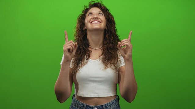Young hispanic woman smiling pointing both index fingers upward while looking up in green studio; optimism hope celebration. - Powered by Adobe