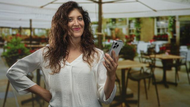 Woman smiling while holding a smartphone on a restaurant terrace outdoors under patio umbrellas; connection happiness leisure.