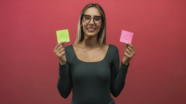 Woman holding pink and green sticky notes with smile and frown in red studio; uncertainty reflection.