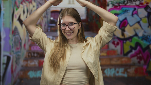 Woman raises arms overhead in graffiti street with vibrant graffiti walls and colorful tags; confidence. - Powered by Adobe