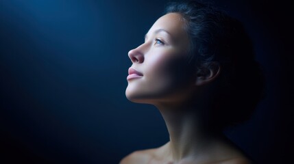 Fototapeta premium Serene profile of a young woman gazing upward in soft natural light, with a tranquil and contemplative expression against a dark background