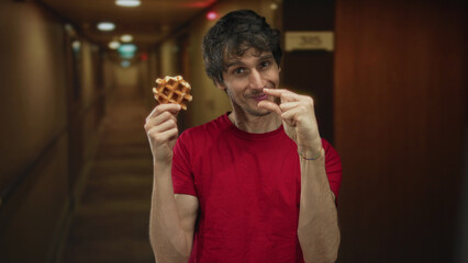 Man smiles while holding a waffle and pinches fingers to show size in a hotel hallway; lighthearted fun.