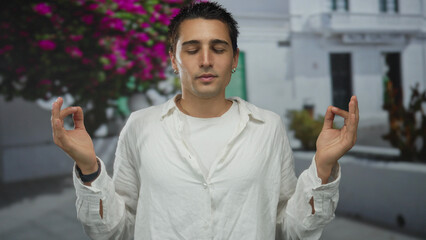 Young man meditating outdoors in a peaceful garden setting with vibrant flowers and background of white buildings