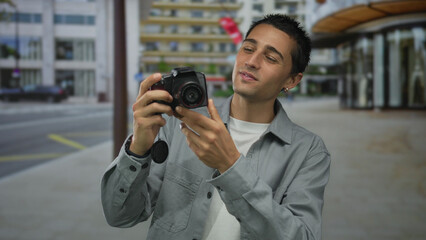 Young hispanic man examines camera with curiosity on an urban city street, surrounded by tall...