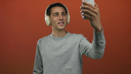 Young hispanic man wearing headphones waves hand while holding smartphone in a studio; virtual connection.