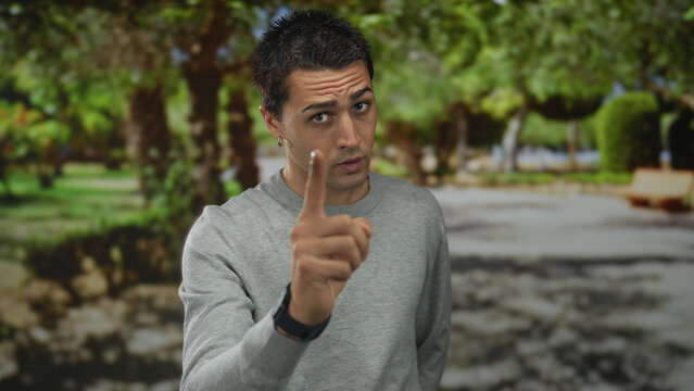 Young hispanic man in grey sweater gesturing seriously with finger pointing in sunny park with green trees and blurred background outdoors during daytime