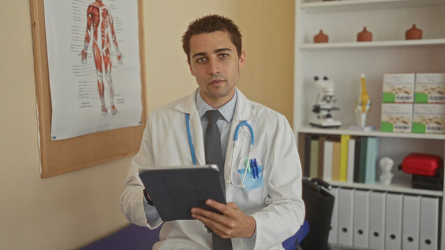 Young hispanic male doctor in a clinic room reviewing data on a tablet with a stethoscope around his neck, surrounded by medical tools and anatomical posters on the wall.