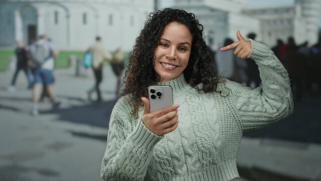 Young hispanic woman taking a selfie with a smartphone outdoors in pisa near the iconic tower on a bustling street filled with tourists.
