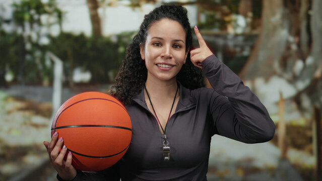Woman holding basketball outdoors touches head playfully, suggesting an idea in a sunny park environment, displaying hispanic heritage and wearing sports attire with a whistle.