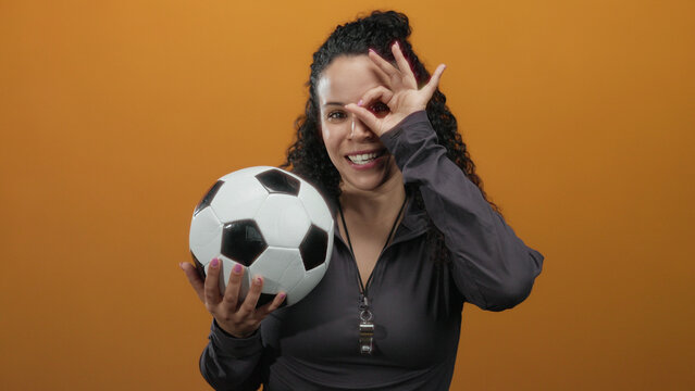 Young hispanic woman making an ok gesture over her eye while holding a soccer ball, standing against an isolated vibrant orange wall background.