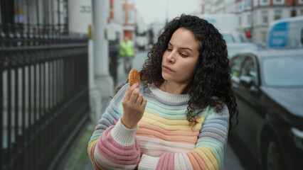 Young woman in colorful sweater examines cookie thoughtfully on busy street with cars passing by in urban outdoor setting reflecting a contemplative mood.