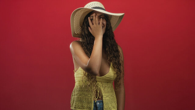 Woman wearing sunhat, hand covering face in studio with yellow crochet top and red backdrop; playful summer laughter. - Powered by Adobe