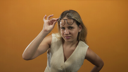 Young blonde woman adjusting round glasses while squinting and leaning forward in orange studio setting; skepticism.