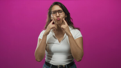 Woman pressing cheeks with both index fingers while puckering lips, wearing glasses and white shirt in pink studio; playful expression.
