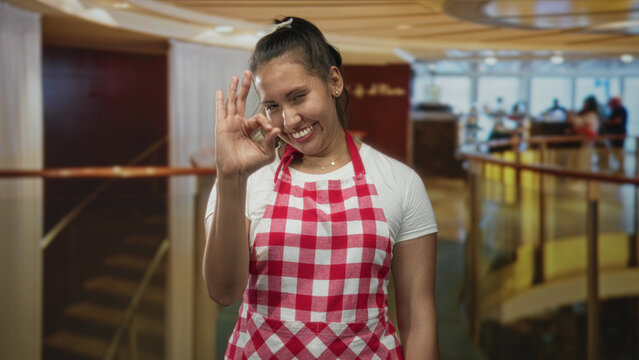 Woman wearing red checked apron makes ok sign with hand in hotel building; cheerful dining service experience.