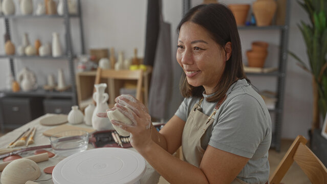 Woman shaping a ball of clay with her hands at a pottery table in a sunlit studio, smiling and wearing an apron; creativity serenity.