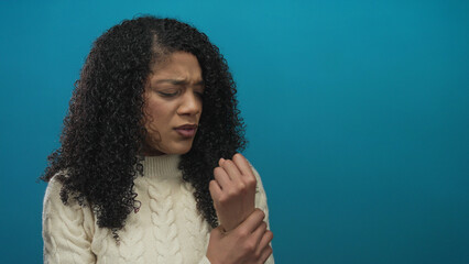 Woman in cream cable knit sweater clutching wrist with furrowed brow under soft lighting in blue...