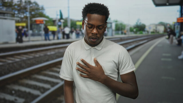 Man wearing glasses clutch chest on railway platform at station building in white polo shirt under a cloudy sky; pain.