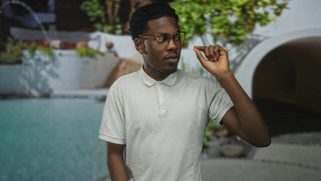 Young african american man pointing finger at sunlit hotel pool outdoors by resort building; serenity. - Powered by Adobe