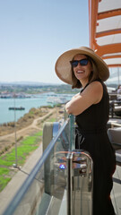 Obraz premium Woman smiling leaning on glass railing on cruise deck overlooking harbor and coastline; serenity vacation.