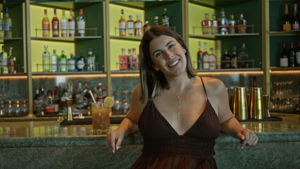 Woman smiling with bare shoulders and cocktail on marble bar counter, looking up while resting hands on edge; relaxed confidence.
