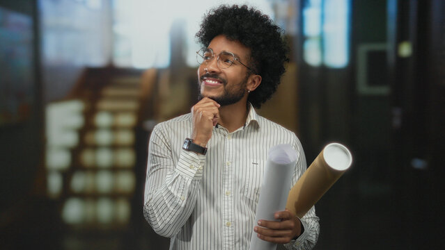 African american man in a hotel holding blueprints indoors, smiling thoughtfully in a modern environment, showcasing creativity and planning in architectural design.