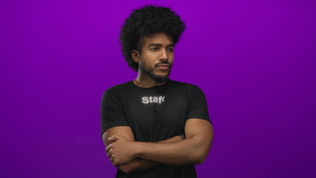 Confident african american man in a staff uniform stands with arms crossed against a vibrant purple wall, exuding professionalism in an isolated studio setting.