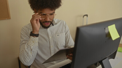 Man working in a modern office wearing glasses and holding documents while focusing on the computer screen with a serious expression in a professional indoor workspace setting.