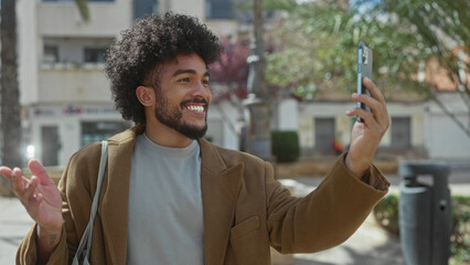 African american man smiling and making a video call in a sunny park with lush greenery, wearing a casual jacket, capturing vibrant outdoor moments with a smartphone.