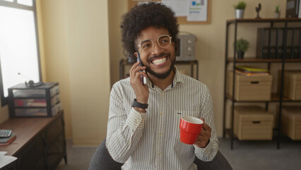 Man talking on phone in indoor office holding red mug smiling with open window and shelves with storage boxes in background.