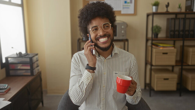 Man talking on phone in indoor office holding red mug smiling with open window and shelves with storage boxes in background. - Powered by Adobe