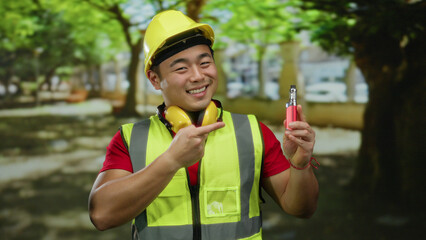 Young man in outdoor park wearing safety gear and hard hat holds electronic cigarette smiling confidently.