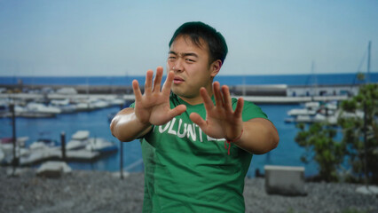 Young man in green shirt labeled volunteer covers face at seaside port with blurred boats under clear blue sky