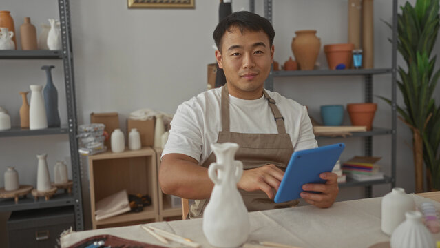 Man holds tablet and taps screen in pottery studio among clay vases and wooden tools on worktable; serenity. - Powered by Adobe
