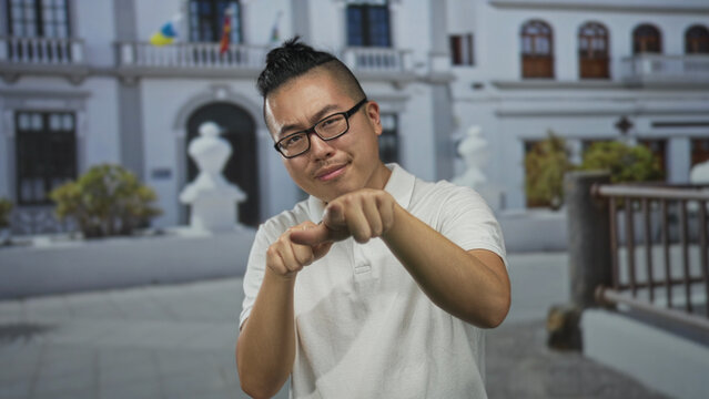 Man wearing glasses and white polo, finger pointing to camera in front of building on a street, pointing finger extended; direct confidence.